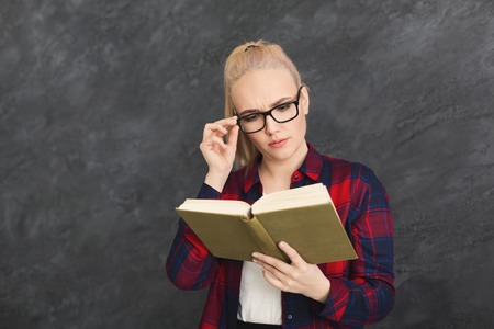 Thoughtful student girl reading book. Young woman in glasses studying at gray studio background, copy spaceの写真素材