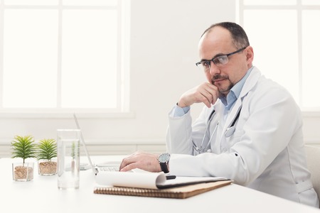 Serious doctor in glasses sitting at desktop, typing on laptop. Physician is working in modern office, medicine concept, copy spaceの写真素材