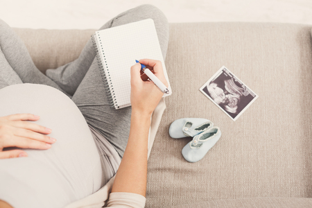Unrecognizable pregnant woman sitting on sofa with blank notebook, pen, tiny shoes and sonogram and writing names for her baby, making shopping list, top view, copy spaceの写真素材