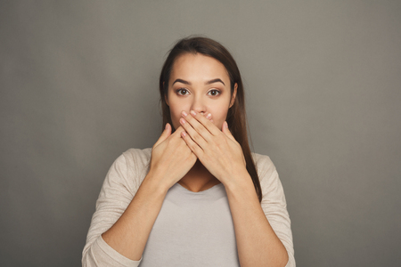 Excited pregnant woman closing her mouth with hands while posing to camera on gray studio background. Asking to keep quiet, speak no evil conceptの写真素材