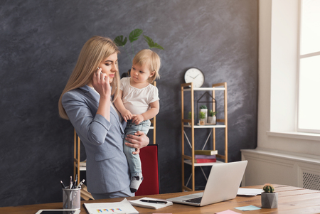 Happy mother working at office with her baby. Young woman talking on phone and using laptop while spending time with her child. Business, motherhood, multitasking and family concept.の写真素材