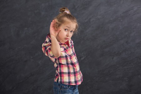 Little girl overhearing gossips. Very curious girl holding her hand near ear, trying to hear something at dark studio background, copy spaceの写真素材