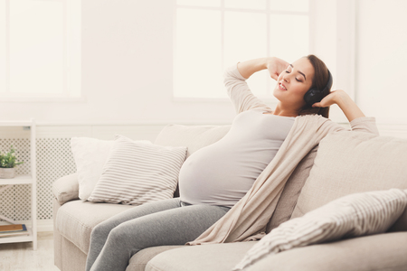 Pregnant woman sitting on sofa and listening music in headphones. Young expectant lady enjoying favorite song home, copy space. Relax, leisure, pregnancy conceptの写真素材