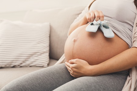 Unrecognizable pregnant woman holding tiny shoes near naked belly. Expectant lady preparing for baby birth, joying maternity, copy spaceの写真素材