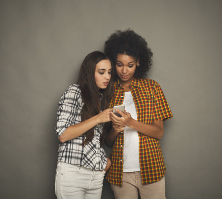 Two interested female friends using smartphone. Shocked women browsing mobile phone, having fun, gray studio background, copy spaceの写真素材