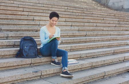Preparing to exams outdoors. Beautiful young female student writing something in notepad sitting on the steps. Education concept, copy spaceの写真素材