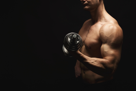 Athletic young man with a dumbbell on a black background. Naked torso, muscular body. Strong chest and shoulder muscles. Studio shot, low key. Bodybuilding concept, copy spaceの写真素材