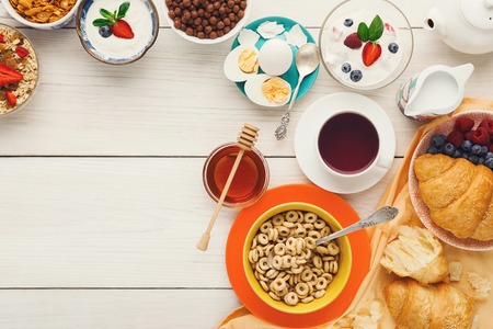 Rich breakfast menu background. French crusty croissants, muesli, yogurt, coffee, berries, honey and boiled eggs for tasty morning meals on wooden table, top view, copy spaceの写真素材