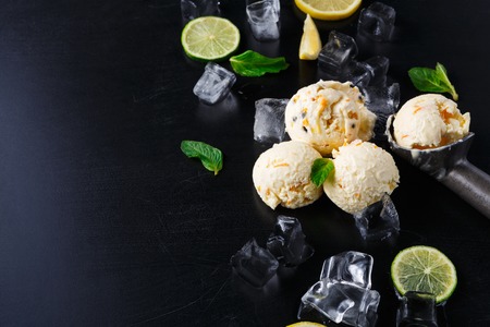 Ice cream scoops with ice cubes and lemon slices and scoop on black background. Delicious cold sweet dessert, top view, copy spaceの写真素材