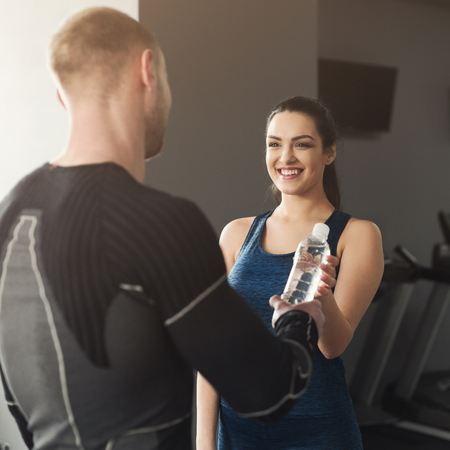 Fitness couple having rest and talking, sharing water at gym. Smiling woman giving plastic bottle to man, talking after training, Healthy lifestyle conceptの写真素材
