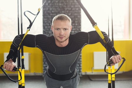 Young man performing TRX suspension training in gym. Muscular guy doing exercise for biceps with elastic rope, copy spaceの写真素材