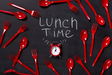 Lunch time background. Red alarm clock and lots of disposable cutlery on blackboard with inscription written with chalk, top view, copy spaceの写真素材