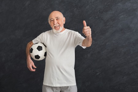 Smiling senior man posing with soccer ball showing thumb up gesture, black background. Hobby, active lifestyle in any age and motivation, copy spaceの写真素材