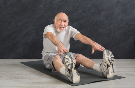 Senior fitness man warmup training indoors. Sporty mature guy makes aerobics exercise, stretching back and legs. Active lifestyle and healthcare in any age, copy spaceの写真素材