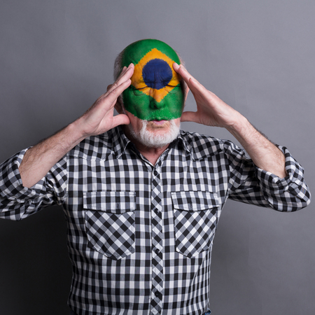 Portrait of sad senior man with flag of Brasil painted on his face. Football or soccer team fan handing head, sport event, faceart and patriotism concept. Studio shot at white background, copy spaceの写真素材