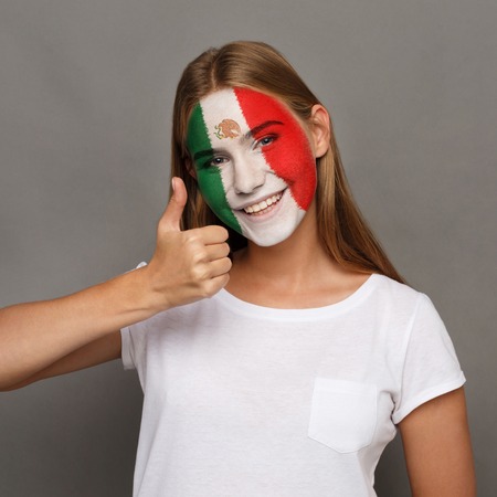 Face of young happy woman painted with flag of Mexica. Football or soccer team fan, sport event, faceart and patriotism concept. Studio shot at gray background, copy spaceの写真素材
