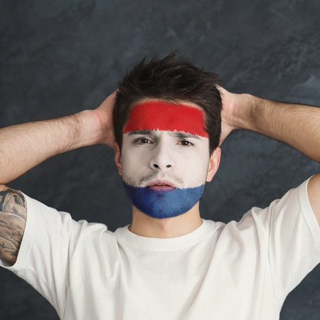 Face of young man painted with flag of Holland. Football or soccer team fan, sport event, faceart and patriotism concept. Studio shot at gray background, copy spaceの写真素材