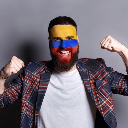Face of young happy bearded man painted with flag of Columbia. Football or soccer team fan, sport event, faceart and patriotism concept. Studio shot at gray background, copy spaceの写真素材