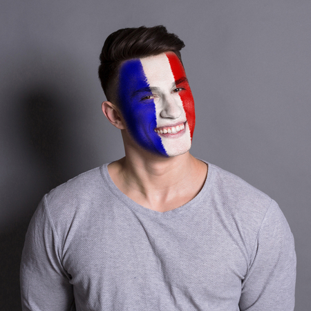 Face of young happy man painted with flag of France. Football or soccer team fan, sport event, faceart and patriotism concept. Studio shot at gray background, copy spaceの写真素材