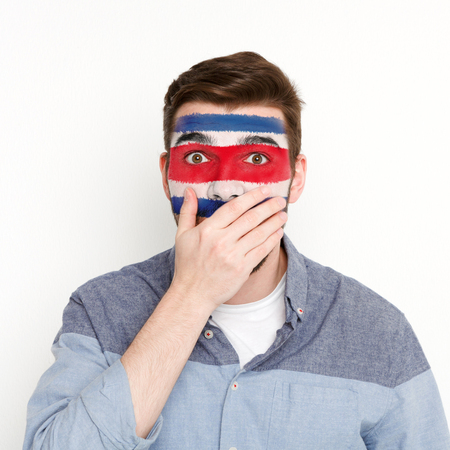 Face of young surprised man painted with flag of Costa Rica. Football or soccer team fan, sport event, faceart and patriotism concept. Studio shot at white background, copy spaceの写真素材