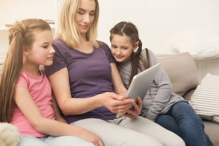 Beautiful young woman and her charming little daughters using digital tablet while sitting on sofa at home. Leasure, family conceptの写真素材