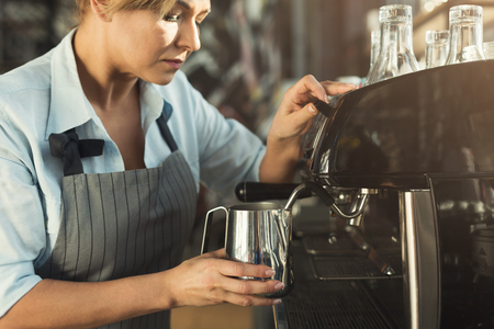 Experienced barista making cappuccino in modern coffee machine. Middle-aged woman steaming milk. Small business and professional coffee brewing conceptの写真素材