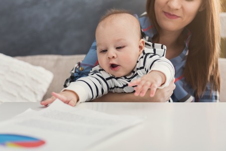 Happy business mom working with diagrams in home office and her cute baby stretching to papers. Business, motherhood, multitasking and family concept, closeupの写真素材