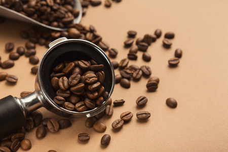 Filter holder with ground coffee, metal scoop and roasted beans scattered around on beige background...