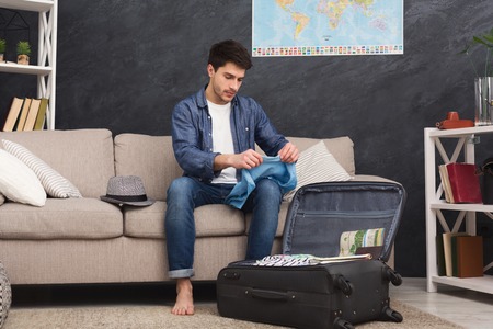 Serious man packing clothes into suitcase, preparing for vacation, sitting on couch at home, copy...