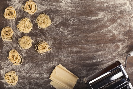 Raw tagliatelle nest and dough slices with pasta machine on kitchen table. Top view on ingredients and equipment for preparing Italian fettuccine, copy spaceの写真素材