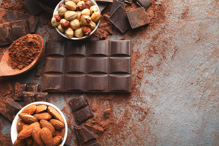 Dark chocolate bar broken in pieces on dark background, cocoa powder and various nuts in bowls closeup, copy space. Sweet wallpaper, confectionery shop advertising and cooking ingredients conceptの写真素材