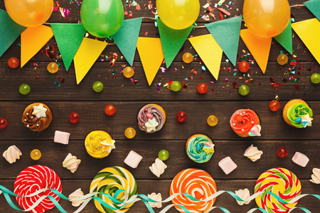 Border of colorful paper flags garland, confetti and three spiral lolipops on rustic wood. Birthday party decoration background, top view, copy space. Mockup for greeting cardの写真素材