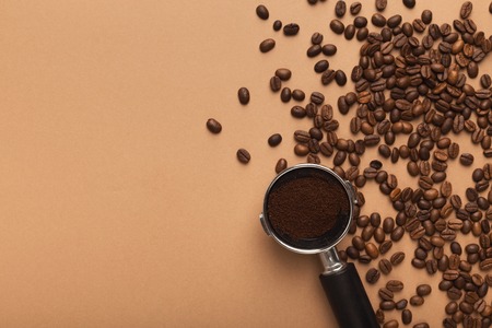 Filter holder with ground coffee and roasted beans scattered around on beige background with copy...