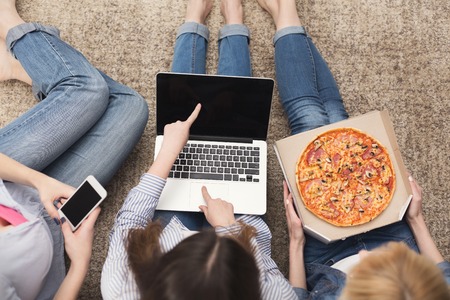 Three girls surfing the web on laptop computer with blank screen and eating pizza at home, top view. Friendship, technology, internet, fast food conceptの写真素材