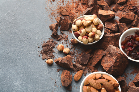 Dark chocolate bar broken in pieces on dark background, cocoa powder and various nuts in bowls closeup, copy space. Sweet wallpaper, confectionery shop advertising and cooking ingredients conceptの写真素材