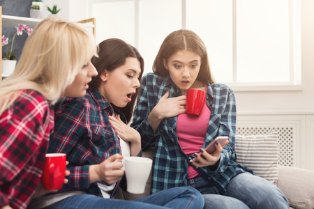 Three shocked female friends with coffee cups looking on smartphone and reading horrifying news. Friendship, shock, news, gossiping, party conceptの写真素材