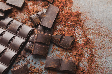 Dark chocolate bar broken in pieces on dark background and cocoa powder closeup, copy space. Sweet...