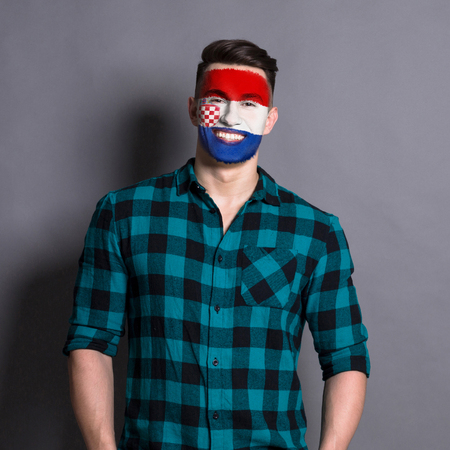Face of young happy man painted with flag of Croatia. Football or soccer team fan, sport event, faceart and patriotism concept. Studio shot at gray background, copy spaceの写真素材