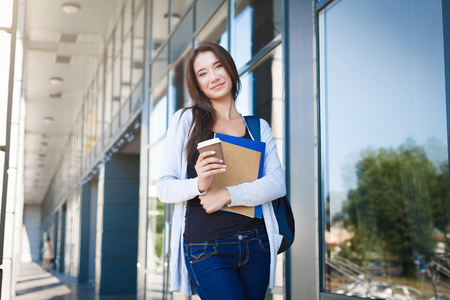 Young female student, with books and backpack. Staying outside before lesson and drinking coffee. Thinking about upcoming day. Lifestyle and education conceptの写真素材