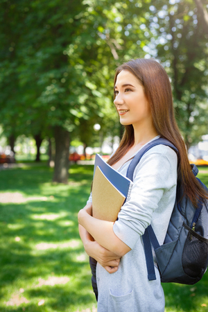 Young female asian student, with books and backpack. Staying in the park after lesson and smiling. Thinking about upcoming day. Lifestyle and education conceptの写真素材