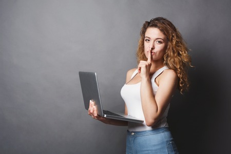 Young woman standing with laptop and showing hush sign. Redhead girl holding open notebook in her hand and work on it. Information, technology concept, copy spaceの写真素材