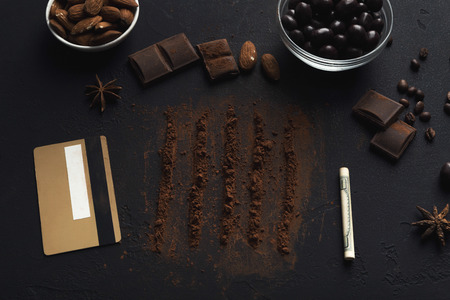 Conceptual chocolate addiction background. Cocoa lines, credit card, rolled dollar pipe and various candies bowls on black tableの写真素材