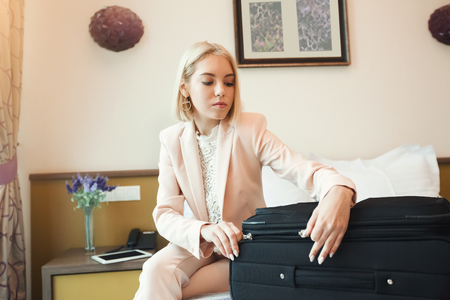 Beautiful young businesswoman in suit unpacking her suitcase while sitting on bed in room. Business lady went to business trip and stayed at hotel, copy spaceの写真素材
