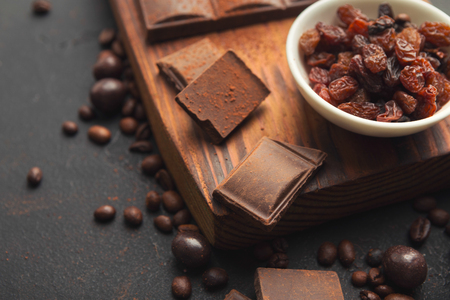 Bitter chocolate bar pieces, raisins bowl and coffee beans on rustic wooden board at black slate background, copy space. Sweet wallpaper, confectionery shop advertising and cooking ingredients conceptの写真素材