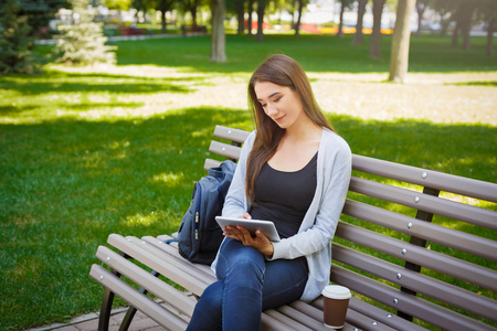 Motivated asian freelancer in the park. Young brunette girl on a bench, drinking coffee and working with tablet online. Lifestyle, technology and remote work conceptの写真素材