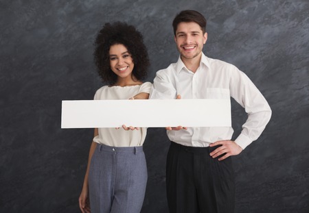 Happy young couple holding white banner with copy space. Man and woman with blank board, gray studio background, copy spaceの写真素材
