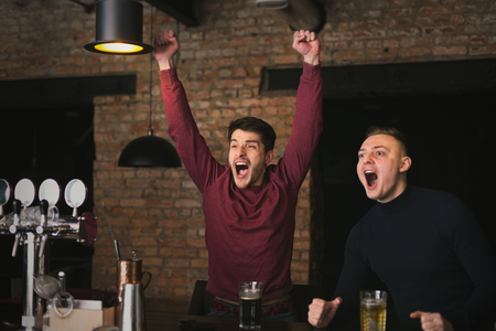 Happy men drinking beer and shouting while watching football match broadcast in bar, copy space. Friends having restの写真素材