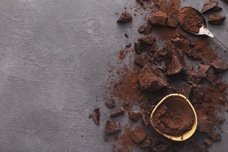 Border of chocolate chunks crushed on dark background and cocoa powder in bowl and spoon, top view,...