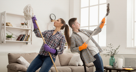 Young couple cleaning home, playing with mop and vacuum cleaner, having fun in living-room....