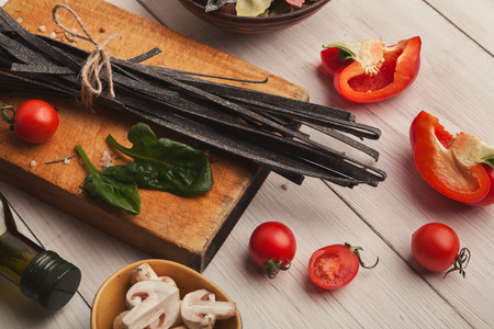 Cooking pasta background. Black colored fettuccine with cuttle fish ink, vegetables and spices on white wooden background, copy space for text or recipeの写真素材
